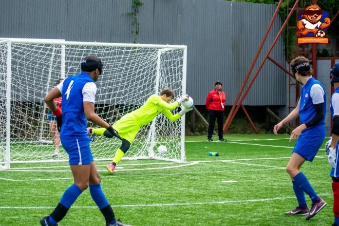 Players wearing blue uniforms and eye shades stand near the goal during a blind football match on an artificial-turf field. A goalkeeper in a bright neon-yellow kit dives across the goalmouth to catch the ball, while other players watch nearby. The goal net, field markings, and a few officials and spectators are visible in the background.
